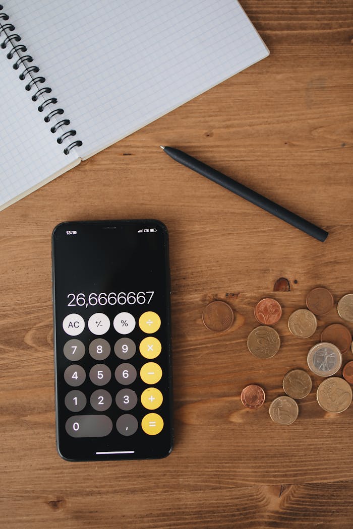 Top view of a smartphone calculator, coins, notebook, and pen on a wooden table for financial concept.